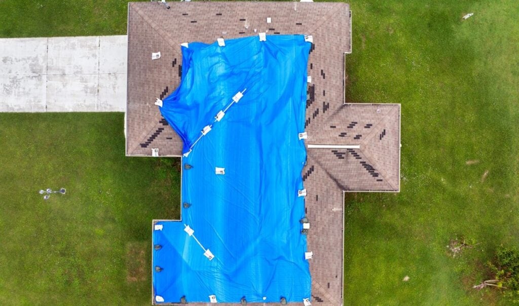Aerial view of a house roof covered with a large blue tarp, possibly for repairs. The tarp is secured with weights and covers the majority of the central roof area. Green grass and a driveway surround the house.