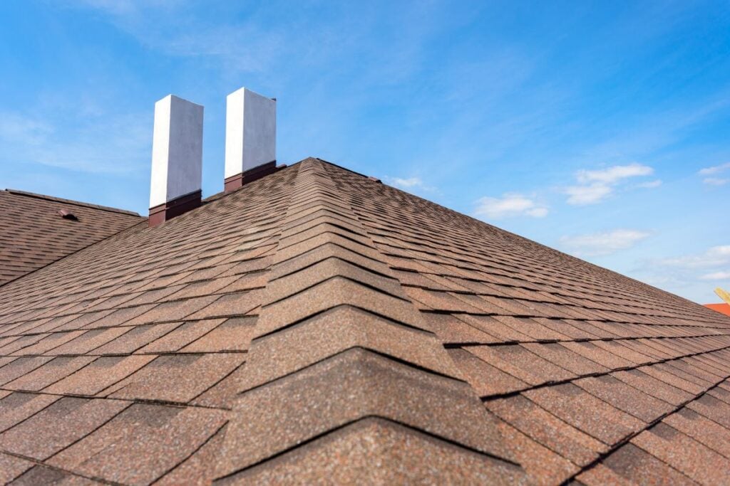 A close-up view of a brown shingled roof with two white chimneys against a blue sky with light clouds.