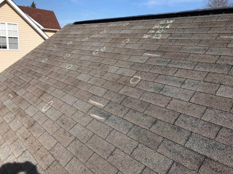 A shingled roof with chalk circles and markings indicating inspection points. Some shingles are marked with lines, likely denoting damage. A house and trees are visible in the background.