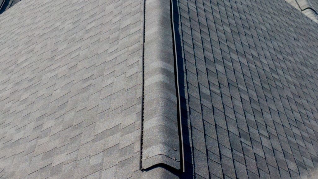 Close-up view of a gray shingled roof, showing the peak with a ridge cap running along the center and symmetrical rows of overlapping shingles on both sides.