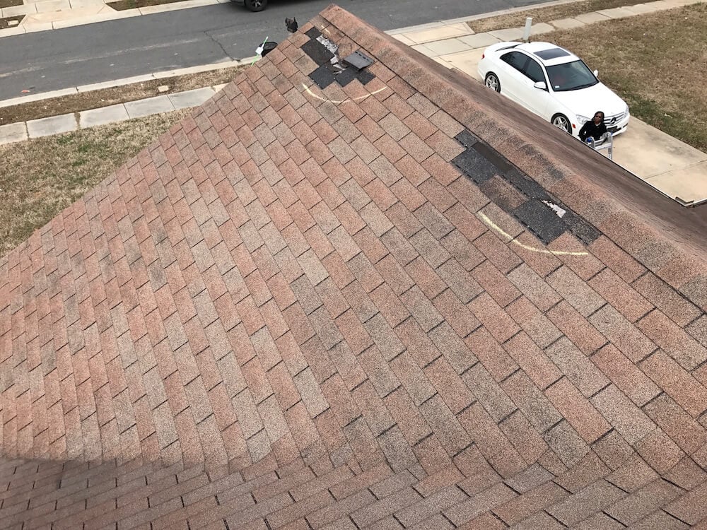 A house roof with missing and damaged shingles, showing areas patched with black shingles. A person and a white car are visible on the street below.