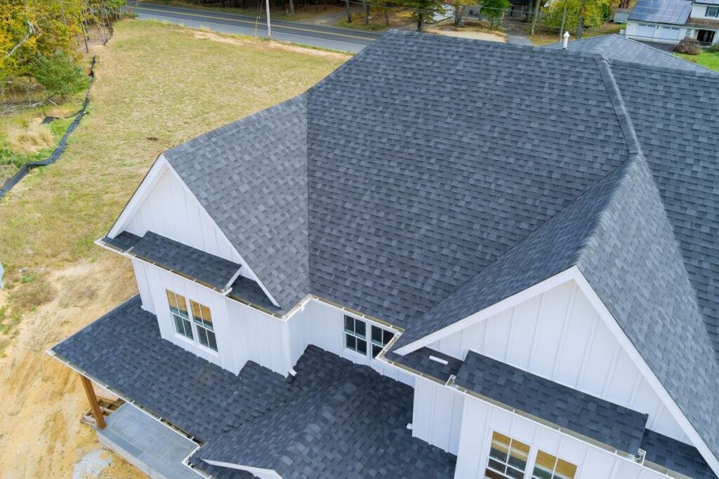 Aerial view of a modern house with dark grey shingle roofing, white siding, and multiple roof angles, situated near a road and an open grassy area.