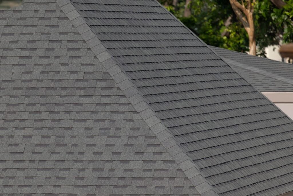 Close-up view of gray asphalt shingle roofs, showing the textured pattern and angles of the rooftops, with some greenery and tree branches visible in the blurred background.