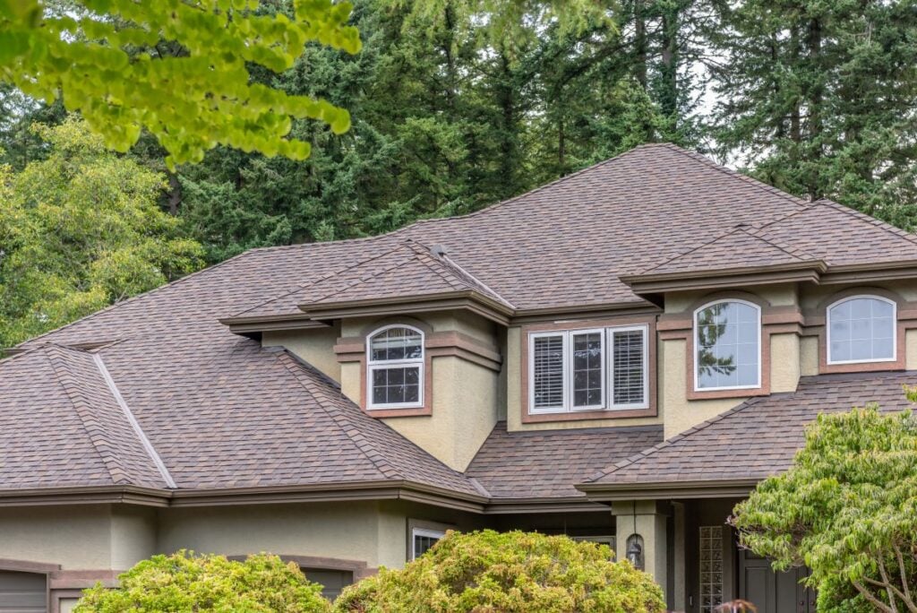 Two-story house with tan walls, brown trim, multiple windows, and a complex dark shingle roof surrounded by green trees and shrubs, seen from an angle showing the upper part of the home.