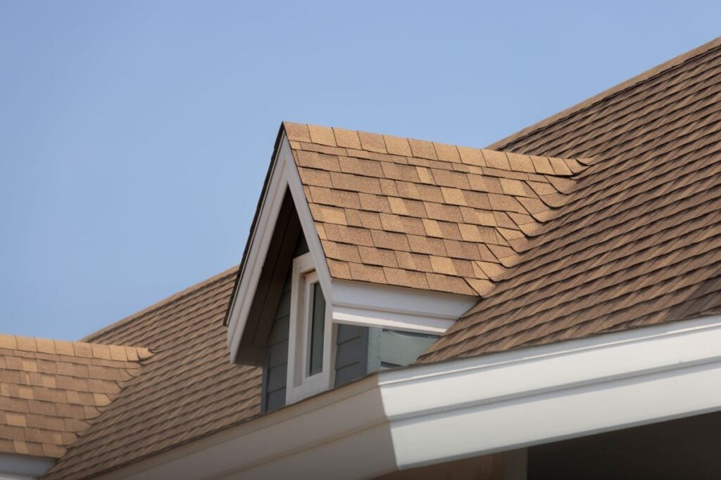 A close-up view of a house roof with brown asphalt shingles, a small dormer window, and white trim, under a clear blue sky.