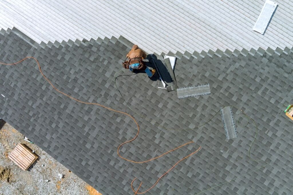 A person installs gray shingles on a roof, with tools and materials scattered nearby. An orange extension cord and green rope are also visible on the dark rooftop.