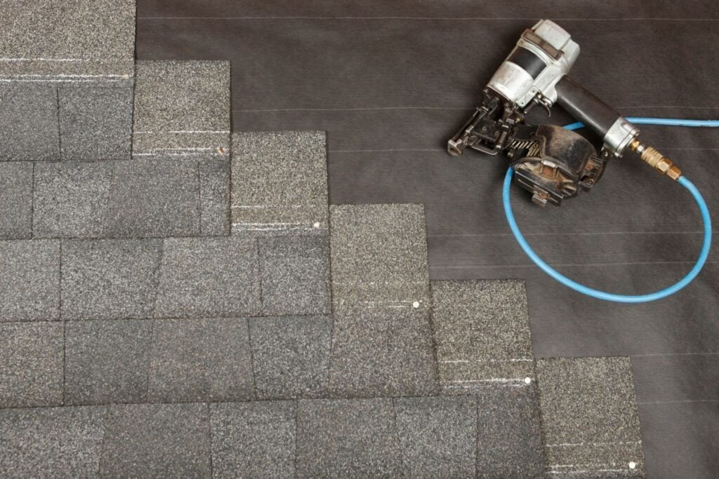 Gray asphalt shingles being installed on a roof, with a nail gun and blue air hose resting next to the partially completed shingles.