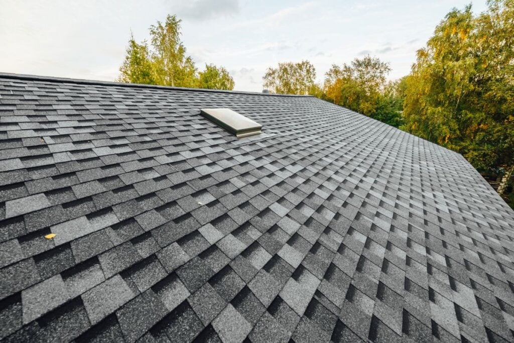A close-up view of a gray asphalt shingle roof with a skylight, surrounded by green trees under a partly cloudy sky.