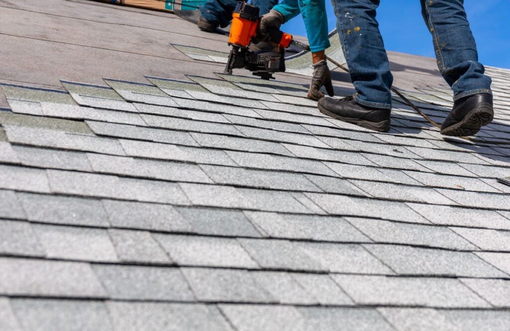 Two workers wearing gloves and jeans use a nail gun to install gray asphalt shingles on a roof under a clear blue sky.