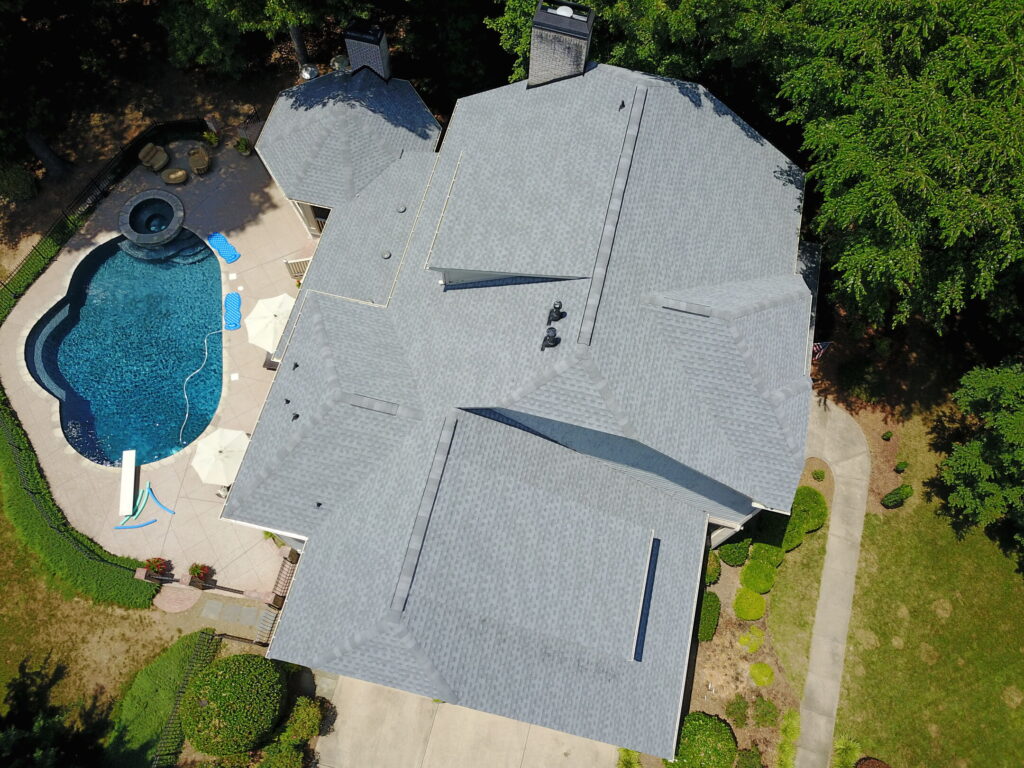Aerial view of a house with a gray shingled roof, a swimming pool, and a surrounding yard with trees.