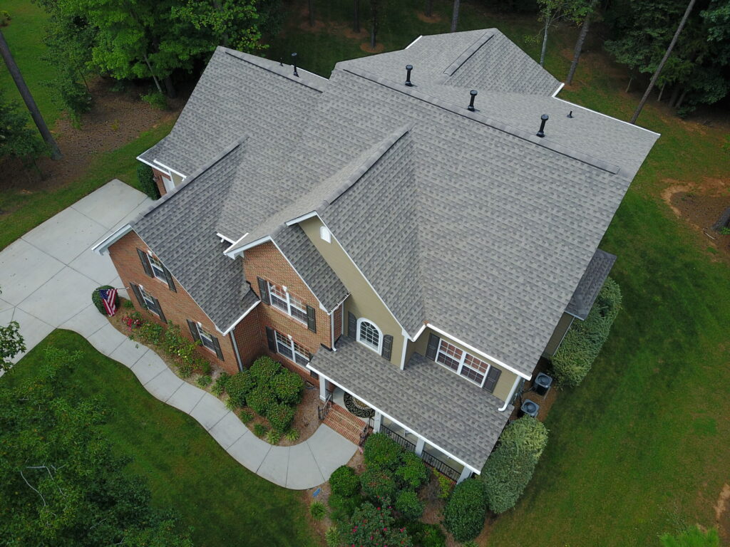 An aerial view of a multi-story brick and tan house with a gray shingled roof, surrounded by a lawn and trees.