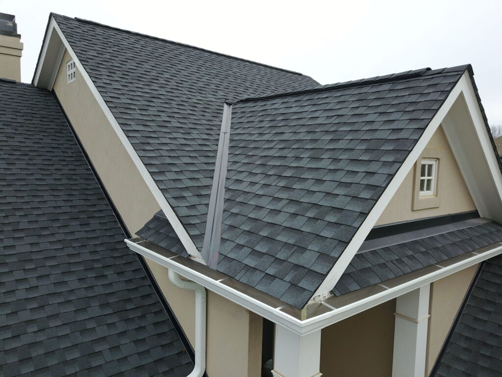 An aerial view of a tan house with a newly installed dark gray asphalt shingle roof, white trim, and white gutters.